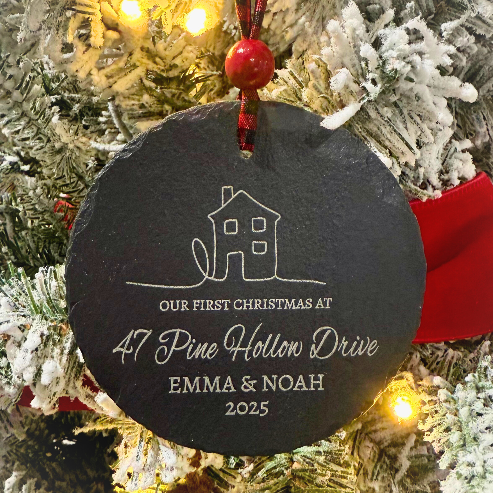 Our First Christmas At personalized 4-inch slate ornament with a cozy house design, customized with couple’s names, street name, and year, hanging from a red and black buffalo plaid ribbon with a red bead.