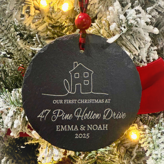 Our First Christmas At personalized 4-inch slate ornament with a cozy house design, customized with couple’s names, street name, and year, hanging from a red and black buffalo plaid ribbon with a red bead.